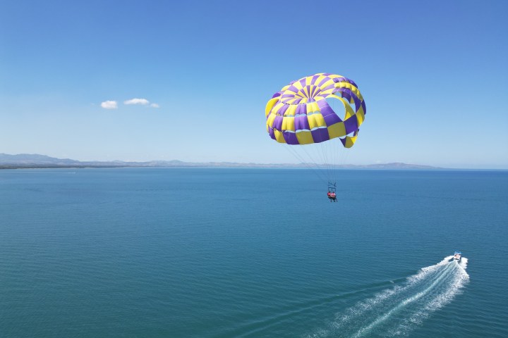 Parasailing over a vast blue ocean with a yellow and purple parachute, pulled by a boat.
